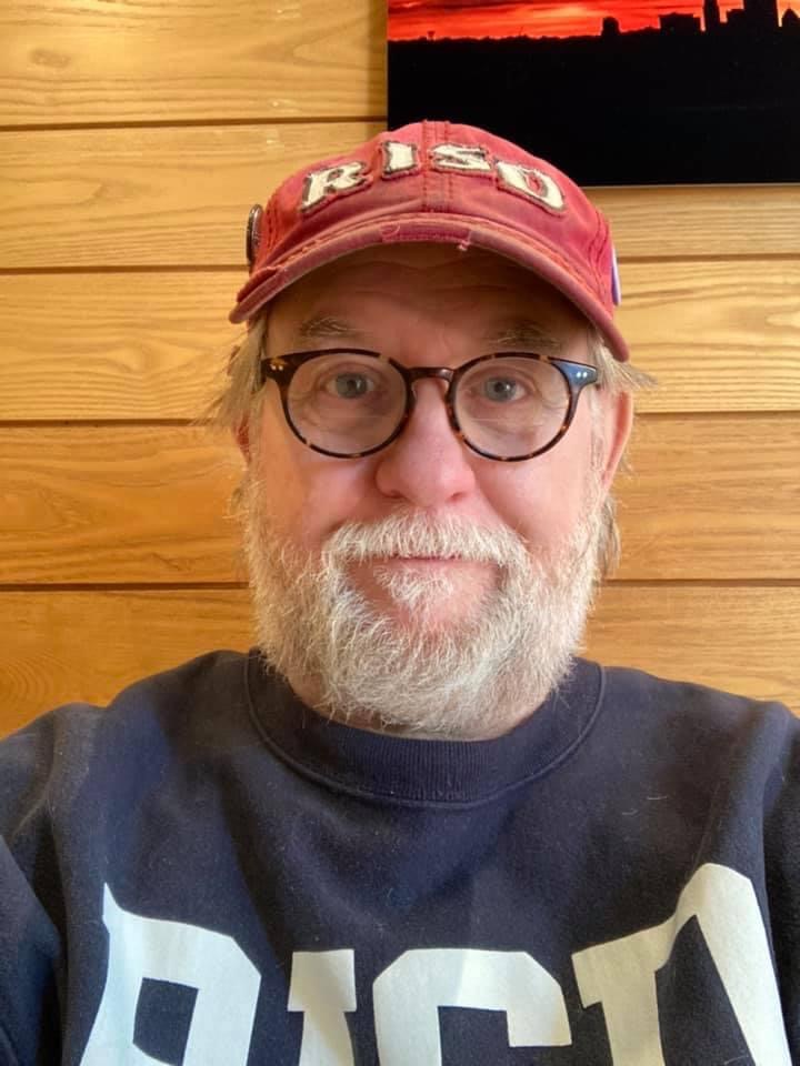 Man wearing a red hat that reads, "R.I.S.D." with a blue sweatshirt on, wearing glasses with a beard looking at the camera.