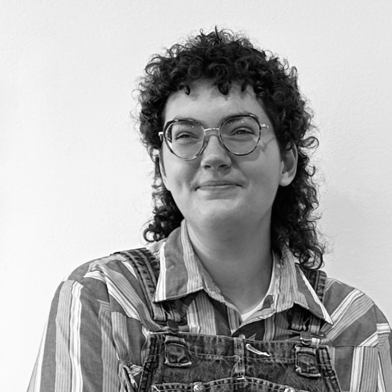 Person with curly hair and glasses wearing a striped shirt with overalls smiling and looking past the camera.