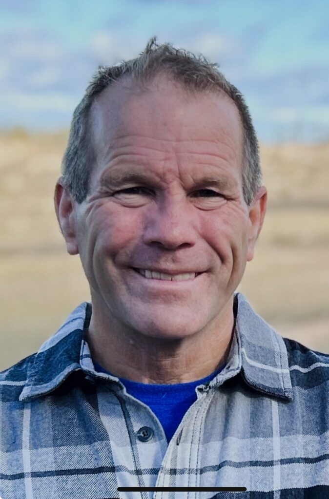 Man in blue and gray plaid shirt over a blue tee, outdoor background.