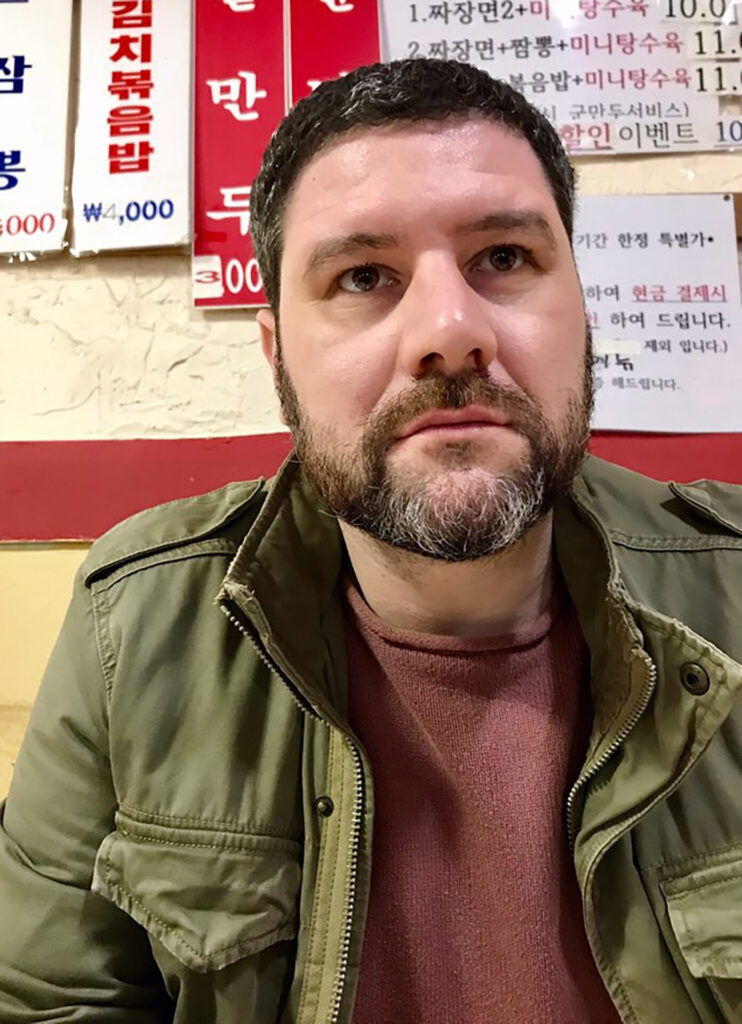 Man wearing green jacket overtop of a maroon shirt sitting in a restaurant.