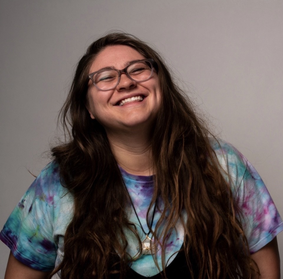 Woman with long brown hair and glasses smiling, wearing a tie-dye shirt with black vest overtop.