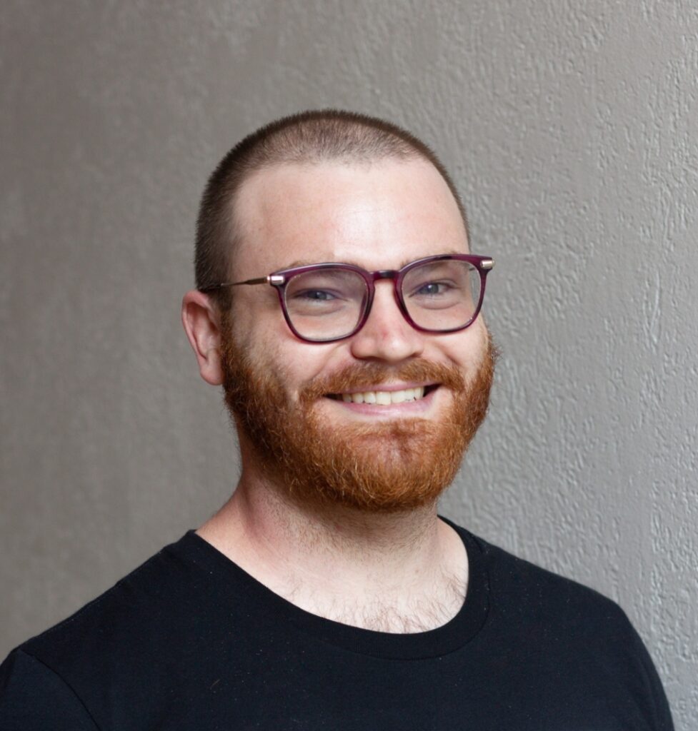 Man with purple glasses and black t-shirt, beard, smiling and looking at the camera.