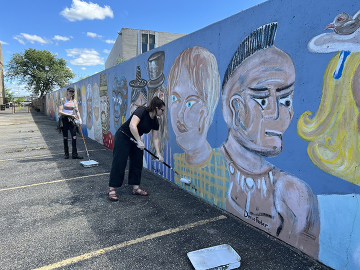 Artists are seen sealing a mural of multiple faces outside in the sun.